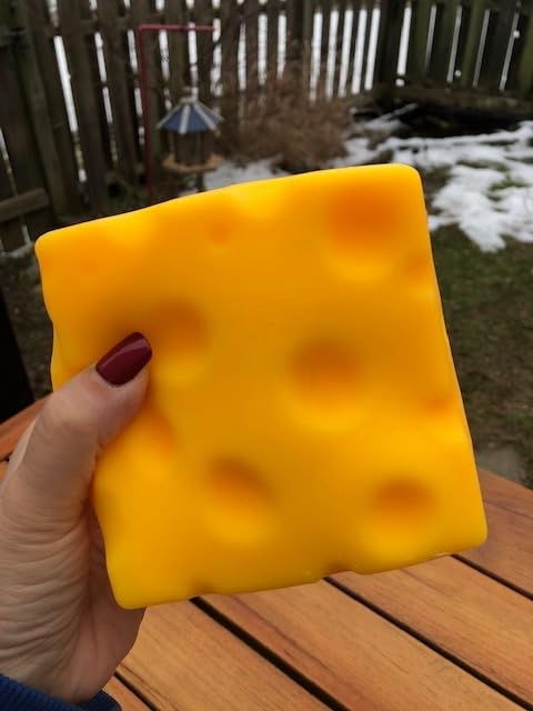 A hand holding the yellow Squeeze Cheese block outdoors, its rounded holes visible across the face