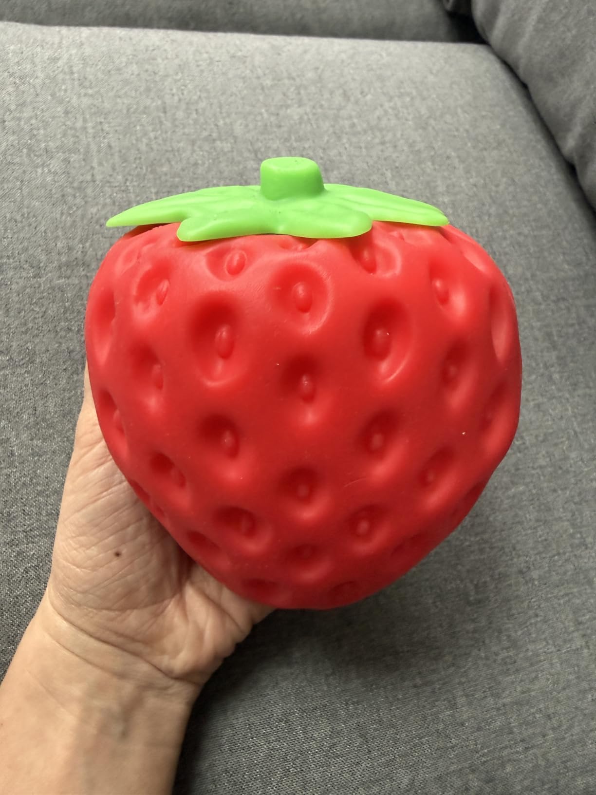 A hand holding the red Squeeze Strawberry on a gray couch, dimpled seed texture and green leaf top filling the palm
