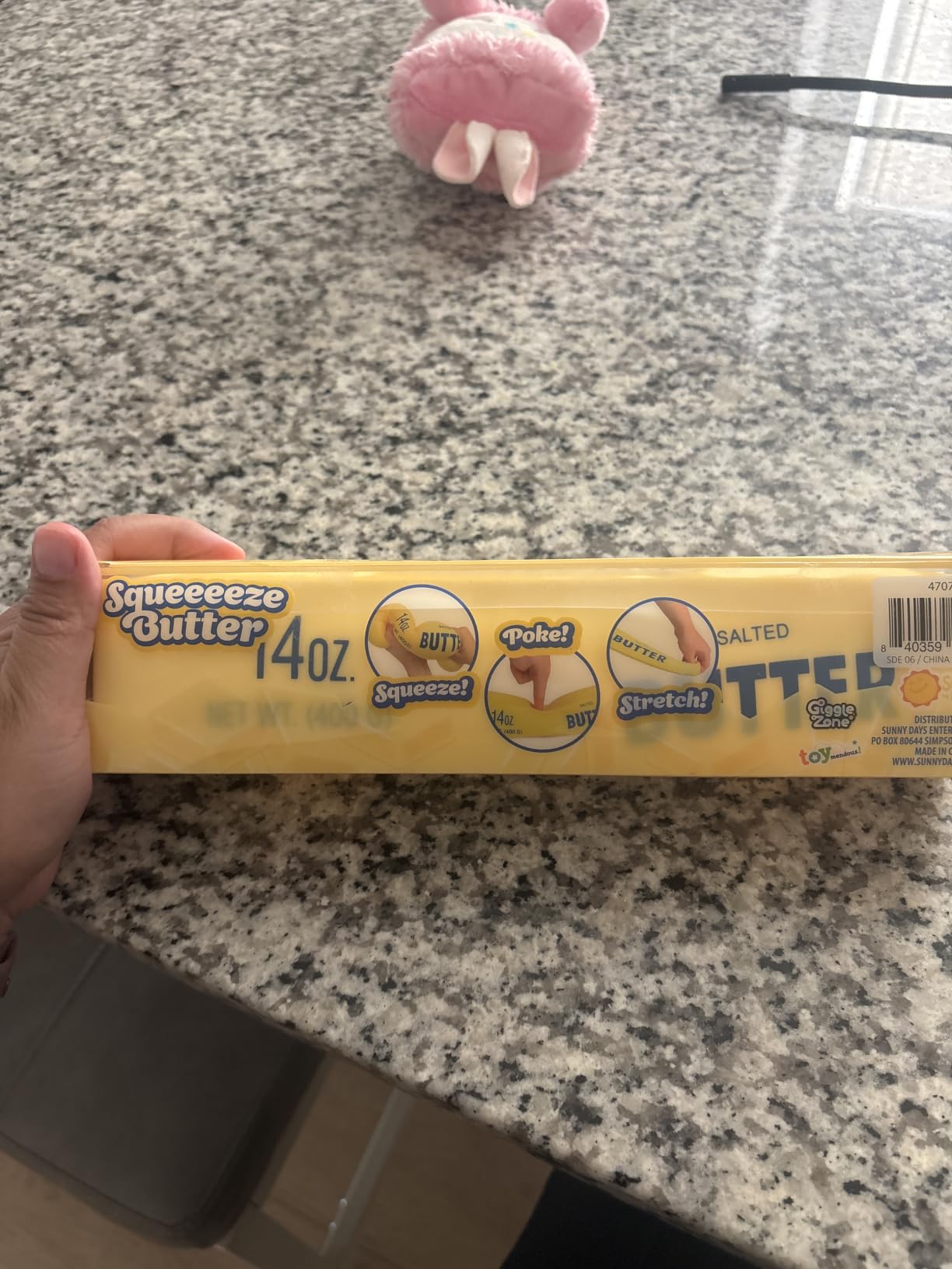 A hand holding the Squeeeeze Butter carton on a granite countertop, with the Squeeze, Poke and Stretch panels along the side